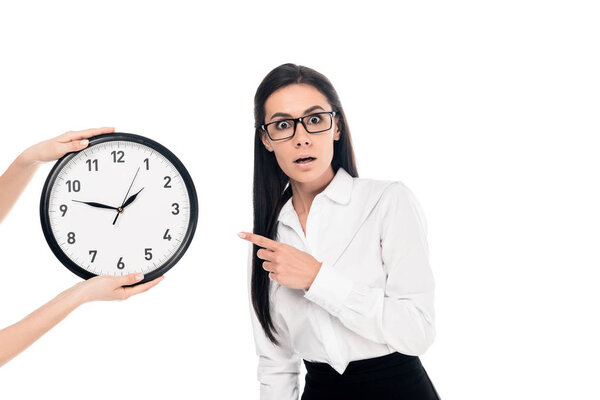 Shocked businesswoman in glasses pointing with finger at clock isolated on white