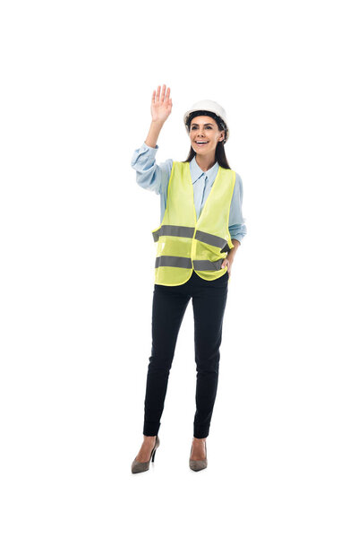 Full length view of smiling engineer in safety vest waving hand isolated on white