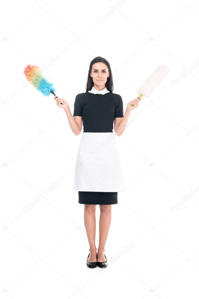Full length view of brunette maid in uniform holding dusters isolated on white