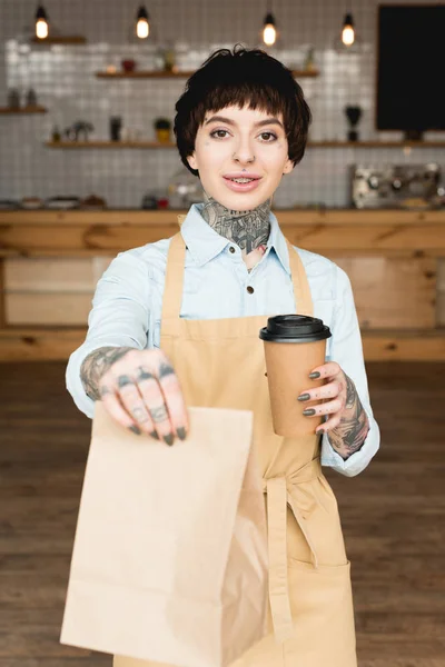 Selective Focus Smiling Waitress Holding Paper Bag Disposable Cup Looking — Stock Photo, Image