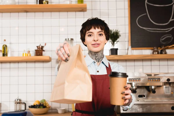 Selektywny Fokus Uśmiechnięta Barista Fartuch Trzymając Worek Papierowy Kubek Jednorazowy — Zdjęcie stockowe