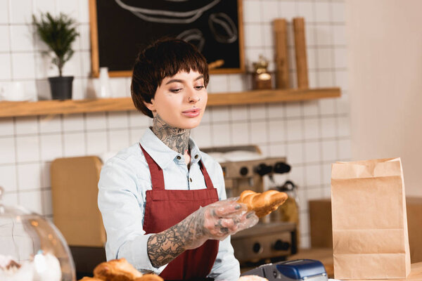 pretty barista in apron holding croissant while standing at bar counter