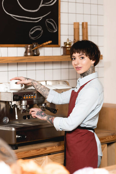 selective focus of smiling barista preparing coffee on coffee machine and looking at camera