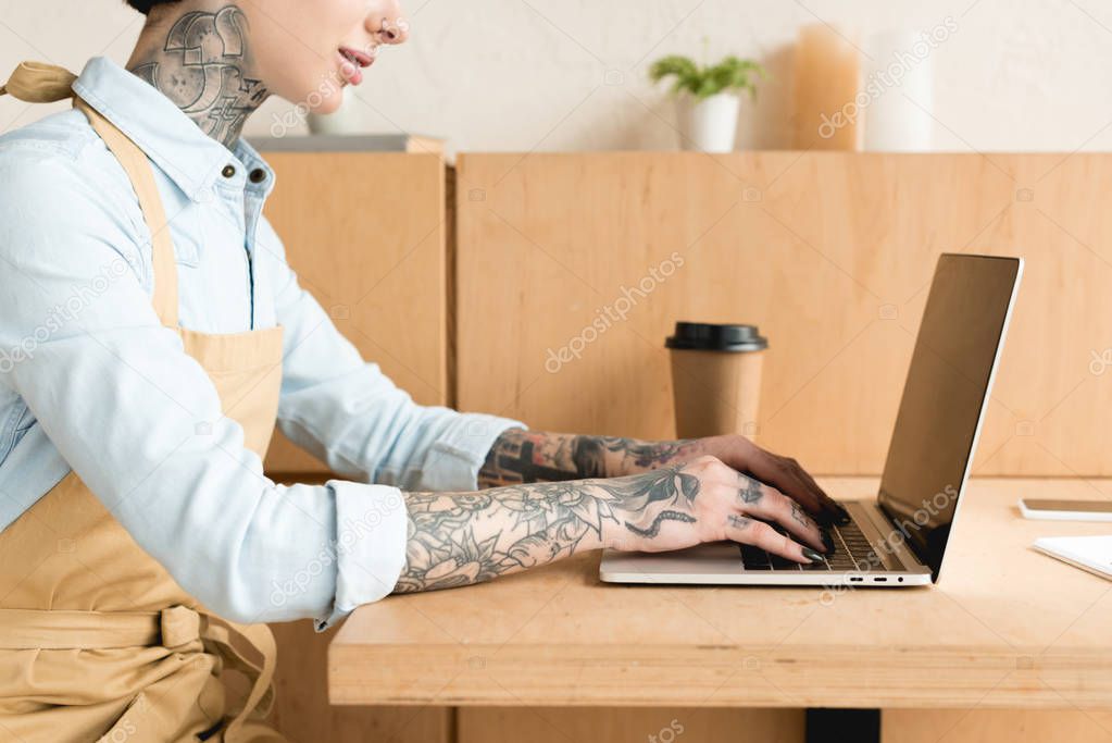 partial view of waitress using laptop while sitting at table near paper cup