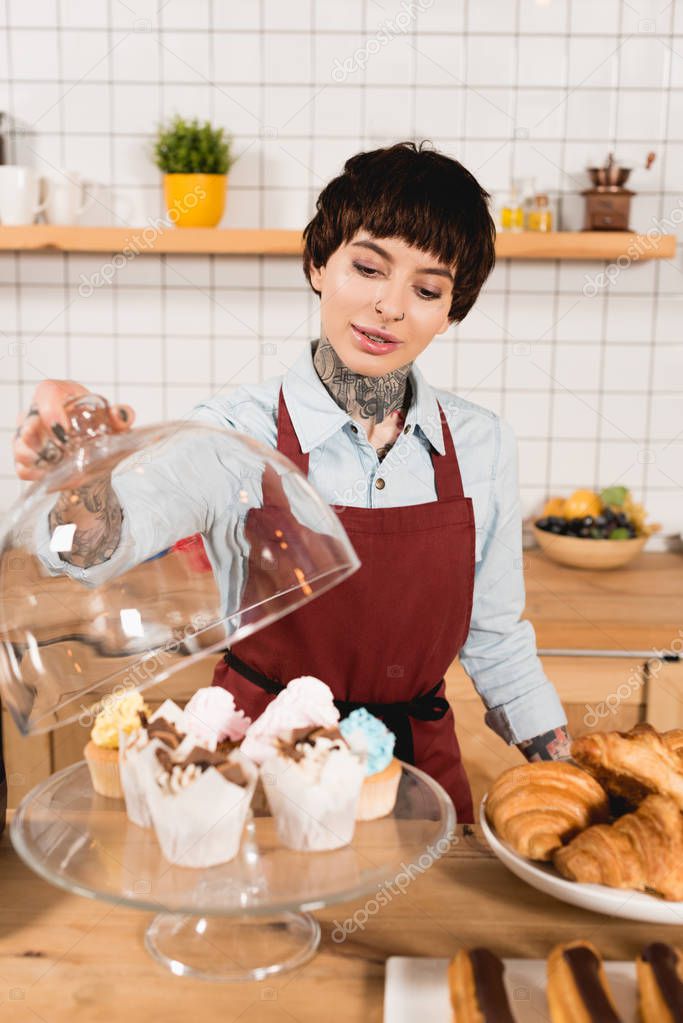 selective focus of pretty barista standing at bar counter with delicious desserts