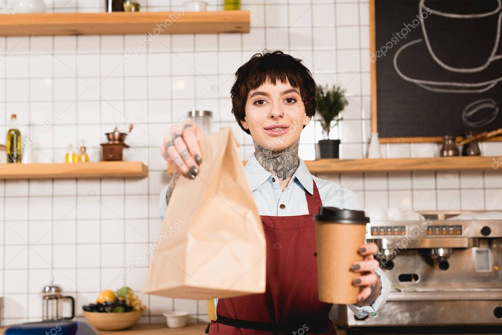 selective focus of smiling barista in apron holding paper bag and disposable cup