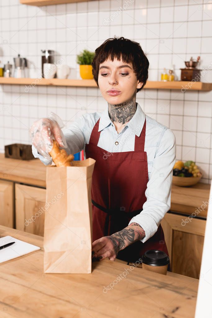 pretty barista in apron putting croissant into paper bag while standing at bar counter