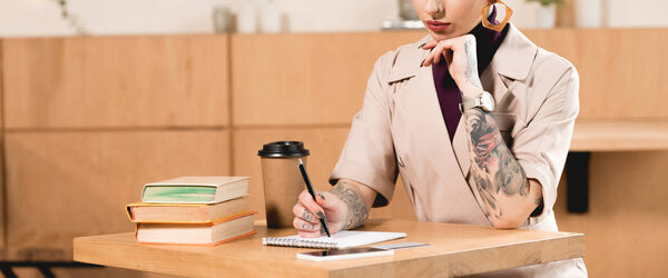 partial view of cafe owner sitting at table in cafe and writing in notebook