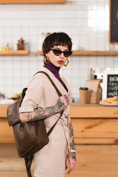 trendy, tattooed businesswoman in sunglasses standing in coffee shop and looking at camera