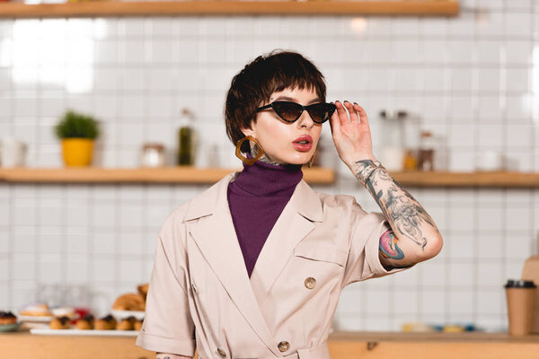 beautiful tattooed businesswoman standing in cafeteria and touching sunglasses