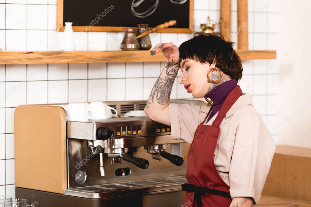 pretty, exhausted barista standing near espresso machine in cafeteria