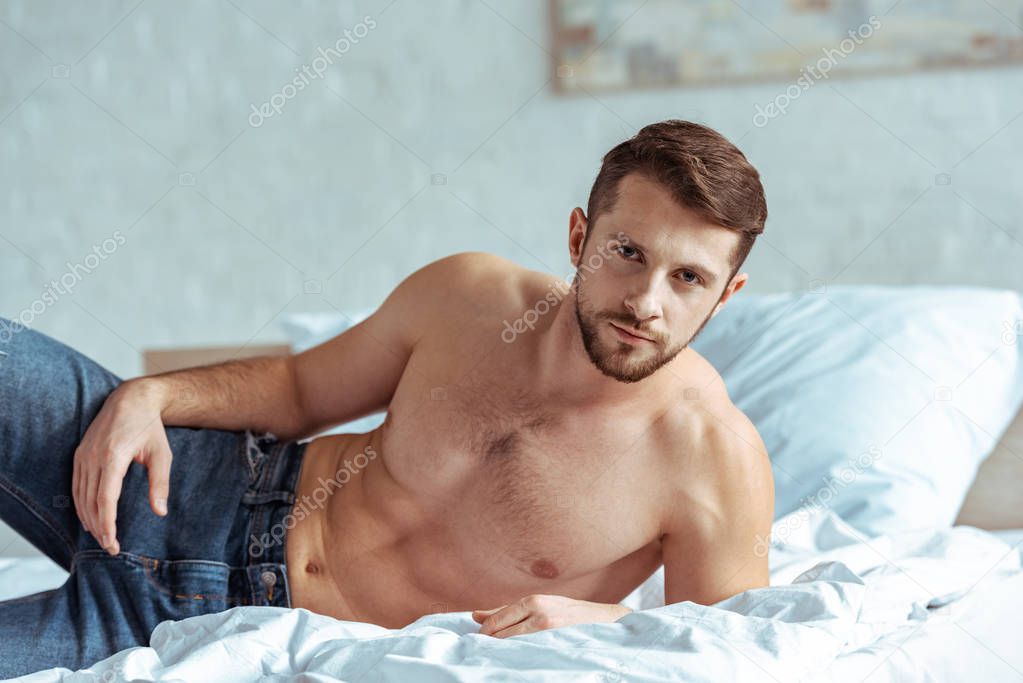 good-looking and muscular man in jeans lying on bed and looking at camera in bedroom 