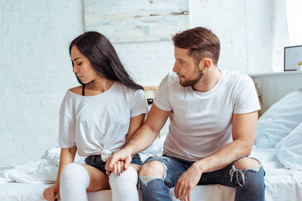 handsome man in jeans holding hand of beautiful and sad woman in t-shirt sitting on bed 