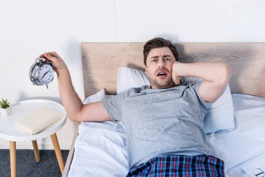 handsome overslept man holding alarm clock while laying on bedding at home clipart