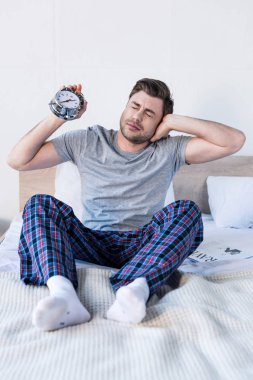 handsome man stretching and holding alarm clock while sitting on bedding  clipart