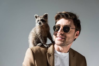 cheerful man in sunglasses with funny raccoon on shoulder on grey clipart
