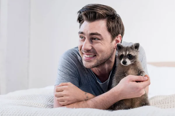 Wesoły Człowiek Przytulanie Adorable Raccoon Łóżku Domu — Zdjęcie stockowe