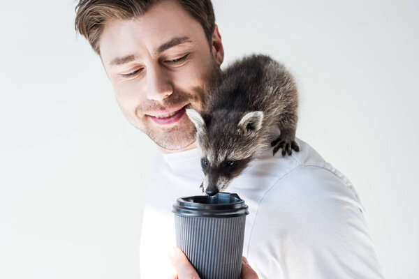 Улыбающийся мужчина с милым енотом пьет из одноразовой чашки на сером

