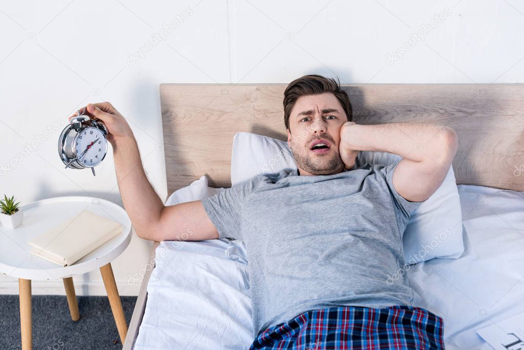handsome overslept man holding alarm clock while laying on bedding at home
