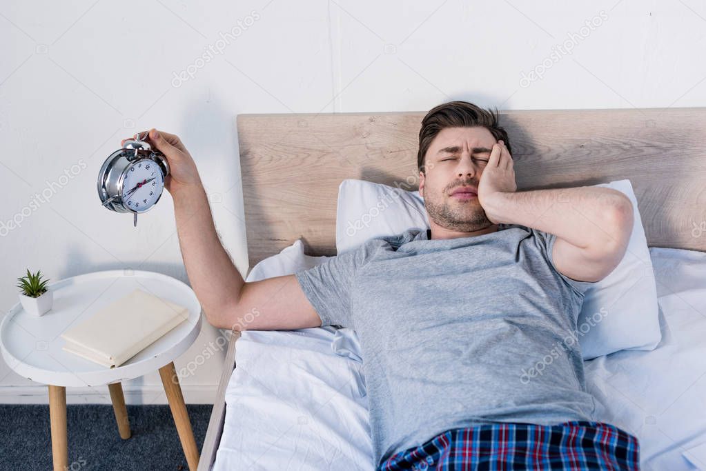 overslept man with closed eyes holding alarm clock while laying on bed