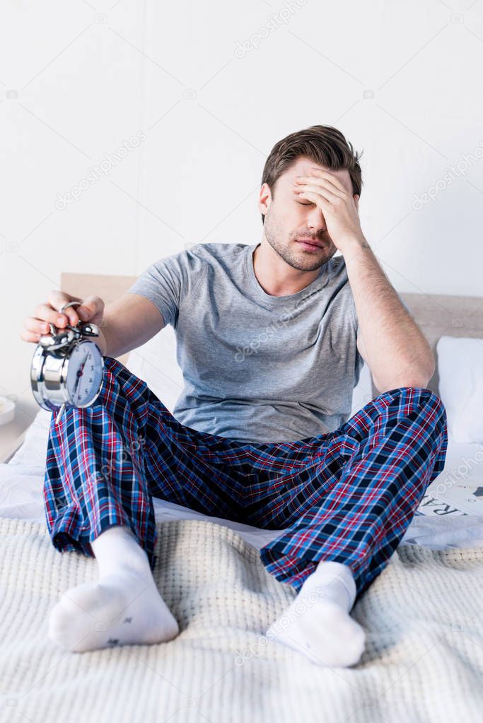handsome overslept man holding alarm clock while sitting on bed at home
