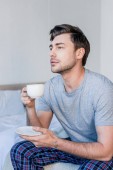pohledný muž sedící na posteli a užívající si ranní kávu