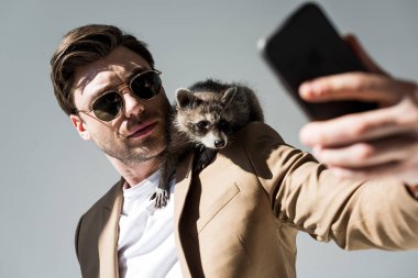 omuz üzerinde sevimli rakun ile yakışıklı adamın seçici odak, gri akıllı telefon ile selfie alarak