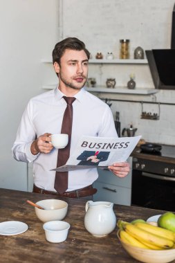 evde mutfakta ayakta iken kendine güvenen adam kahve bardağı ve iş gazetesi tutarak