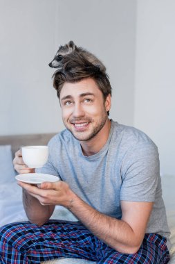 cheerful man with funny raccoon on head holding coffee cup while sitting on bedding clipart