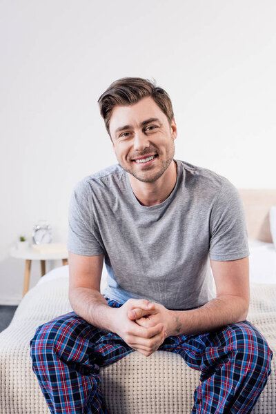 handsome cheerful man sitting on bedding at home and looking at camera