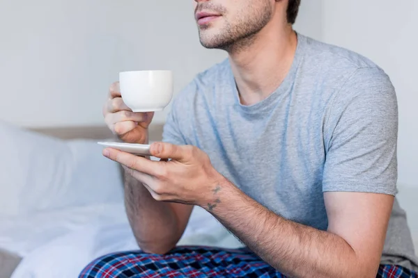 裁剪的看法 男子在灰色的 T恤坐在床上 拿着咖啡杯 — 图库照片