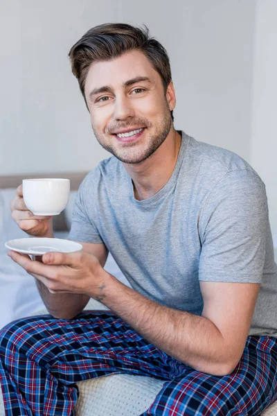 Przystojny Człowiek Uśmiechnięty Piżamie Trzymając Filiżankę Kawy Patrząc Aparat Fotograficzny — Zdjęcie stockowe