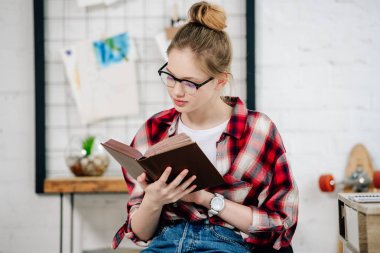 Curious teenage kid in glasses and checkered shirt reading book clipart