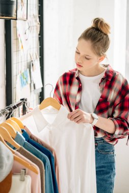 Interested teenage kid in wristwatch holding hanger with white shirt