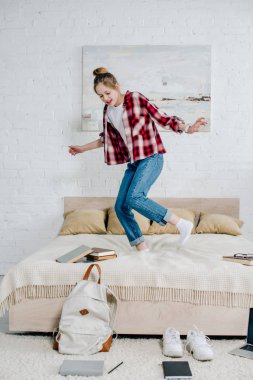 Full length view of smiling teenager kid in jeans jumping on bed at home clipart