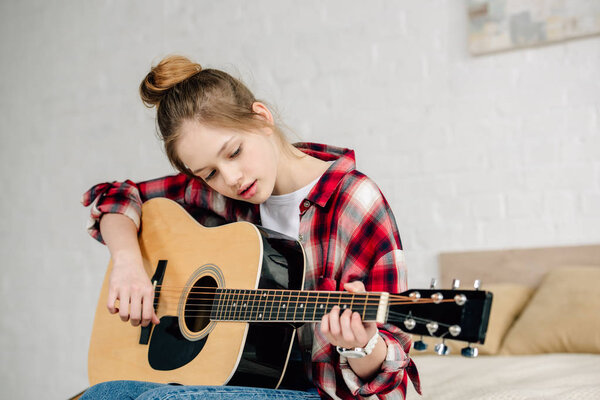 Сконцентрированный подросток в клетчатой рубашке, играющий на акустической гитаре дома
