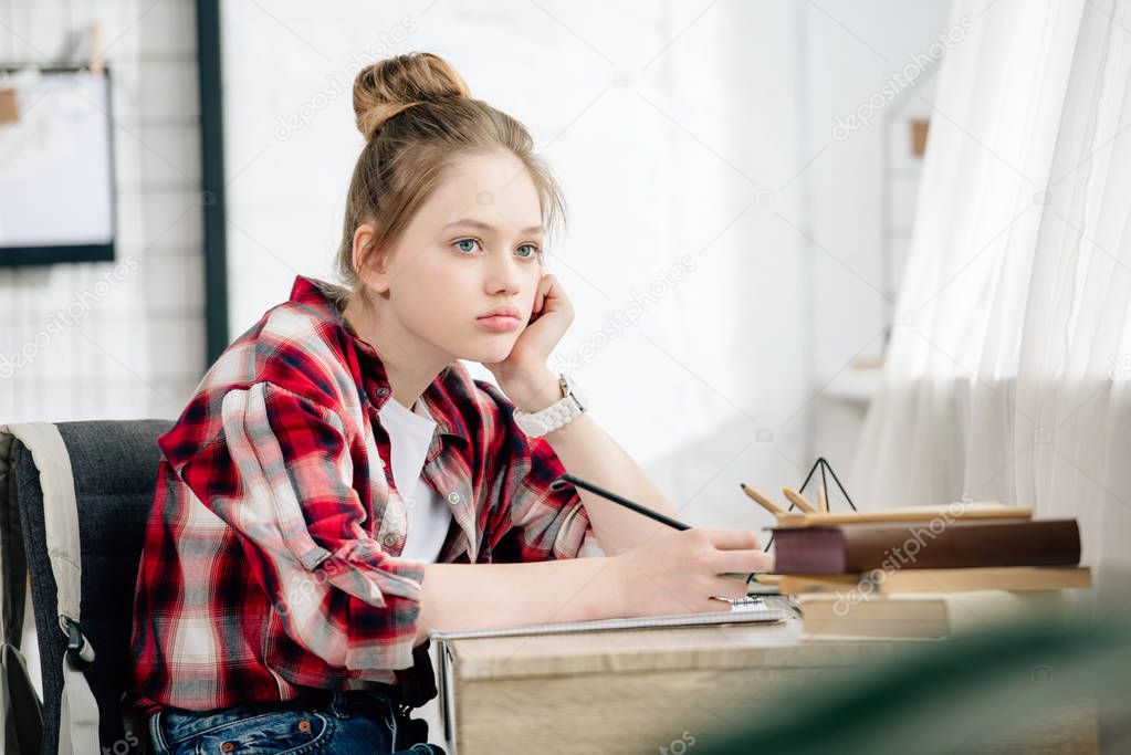 Pensive teenager in checkered shirt holding pen at table while doing homework