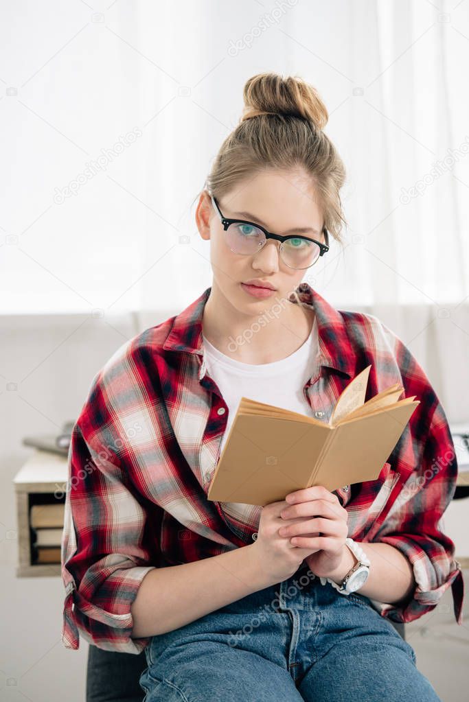 Curious teenage kid in glasses and checkered shirt holding book and looking at camera