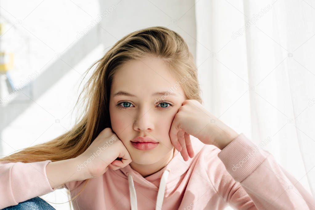 Front view of bored teenage kid in pink hoodie propping face with hands and looking at camera