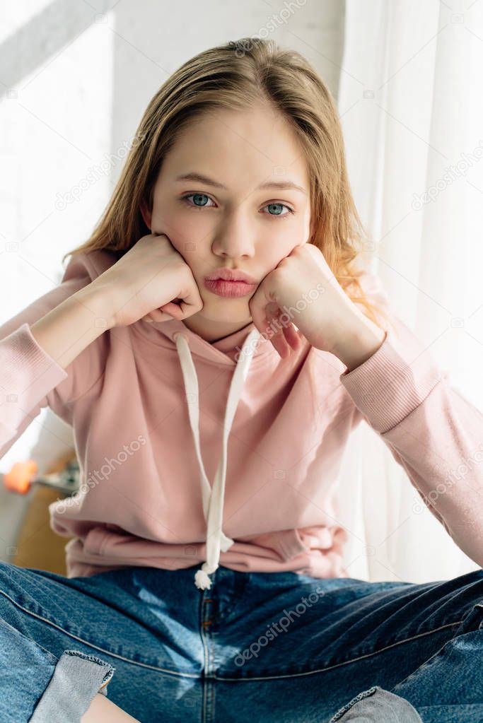 Front view of bored teenage kid in pink hoodie propping face with hands and looking at camera