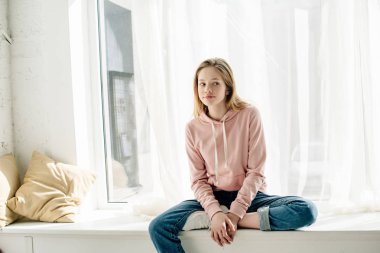 Pensive teenage kid in jeans sitting on window sill and looking away clipart
