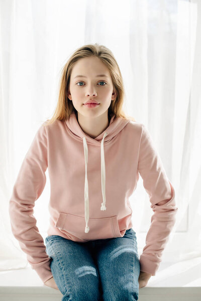 Front view of teenage kid in pink hoodie sitting on window sill and looking at camera