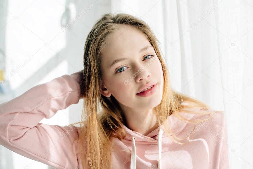 Cute teenage kid in pink hoodie touching hair and looking at camera