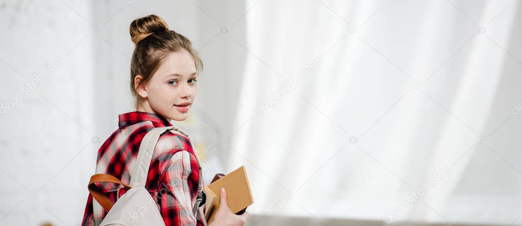 Panoramic shot of teenager with backpack holding books and looking at camera