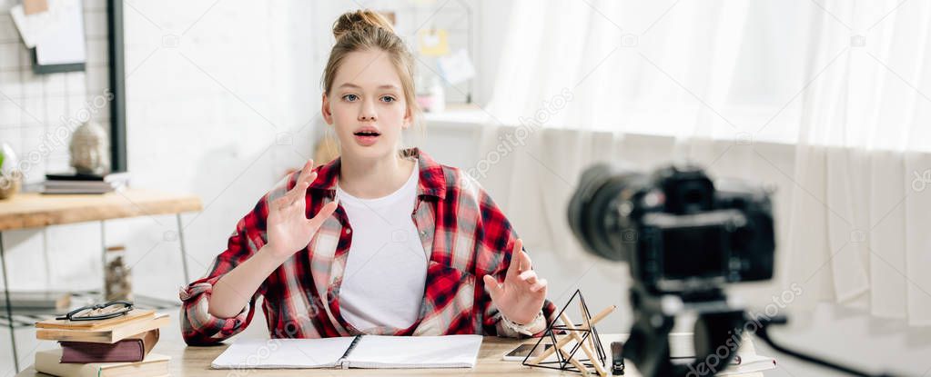 Panoramic shot of teenage blogger in checkered shirt sitting at table in front of video camera