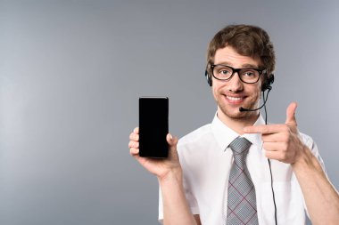 smiling call center operator in glasses and headset pointing with finger at smartphone with blank screen clipart