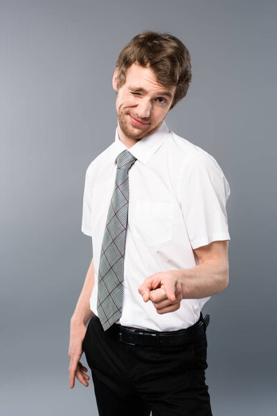 sly businessman pointing with finger at camera and winking on grey background