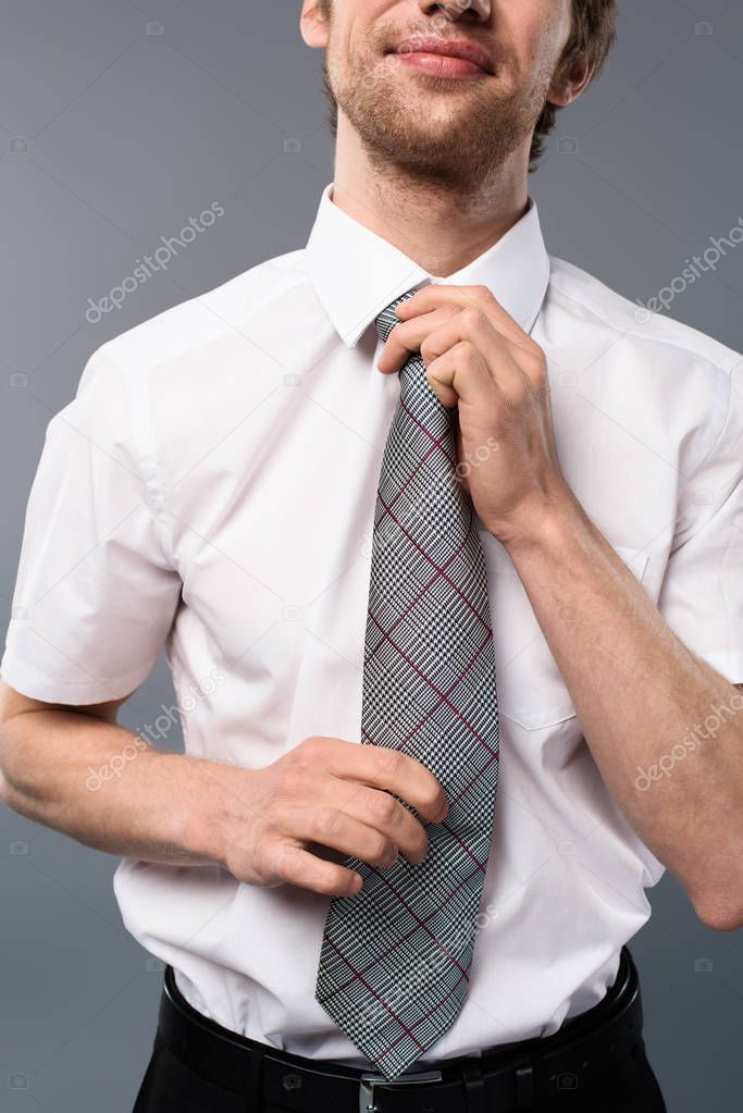 cropped view of cheerful businessman tying tie on grey background