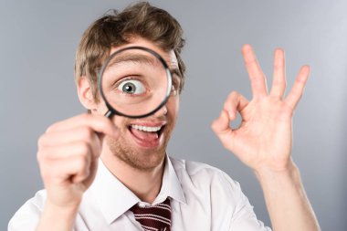 positive businessman with funny face expression looking in magnifier and showing ok sign on grey background clipart