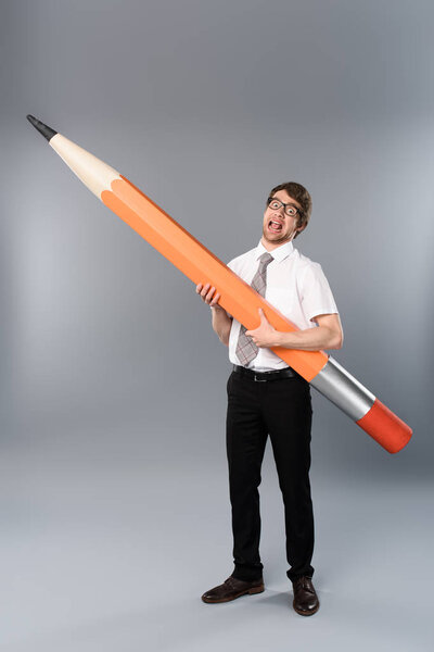 funny businessman in glasses holding huge and heavy decorative pencil on grey background